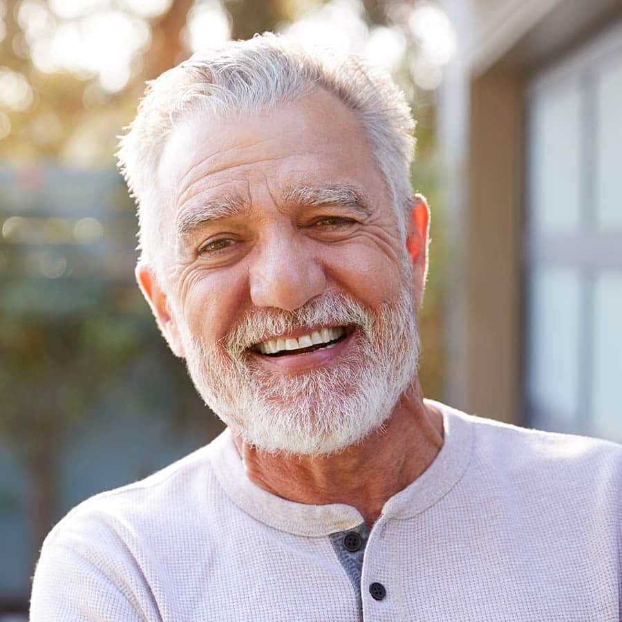An older man with gray hair and a beard smiles warmly outdoors, wearing a light-colored henley shirt. Soft sunlight and greenery are visible in the blurred background.